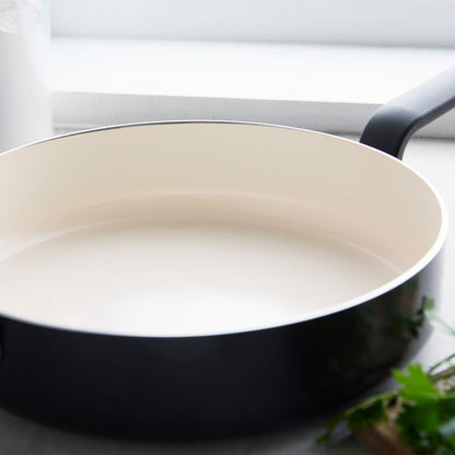 Black ceramic nonstick 5-quart sauté pan with lid on a light kitchen surface, fresh herbs nearby.