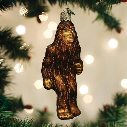A glass ornament shaped like Bigfoot hangs from a Christmas tree branch, surrounded by green pine needles and blurred white holiday lights in the background.