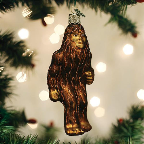 A glass ornament shaped like Bigfoot hangs from a Christmas tree branch, surrounded by green pine needles and blurred white holiday lights in the background.