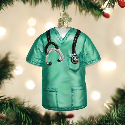 A Christmas tree ornament shaped like turquoise medical scrubs with a stethoscope draped around the neck, hanging on a tree with blurred lights in the background.