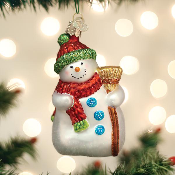 A festive snowman ornament with a red hat, green trim, red scarf, blue buttons, and holding a gold broom hangs on a Christmas tree with soft white lights in the background.