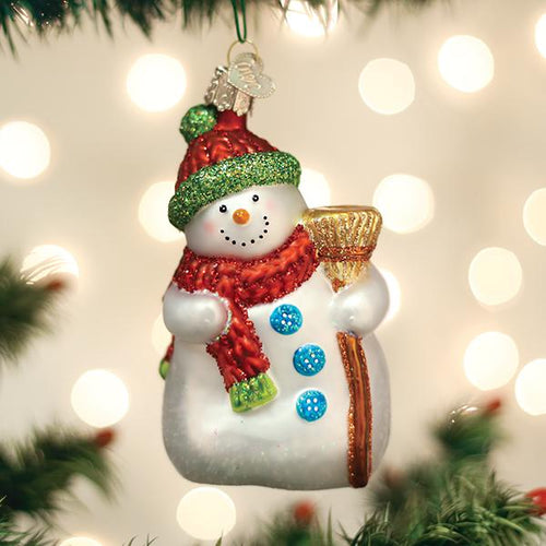 A festive snowman ornament with a red hat, green trim, red scarf, blue buttons, and holding a gold broom hangs on a Christmas tree with soft white lights in the background.