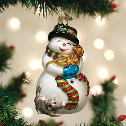 A festive snowman ornament with a red and green scarf holds a golden puppy and has a gray cat by its side, hanging on a Christmas tree with blurred lights in the background.