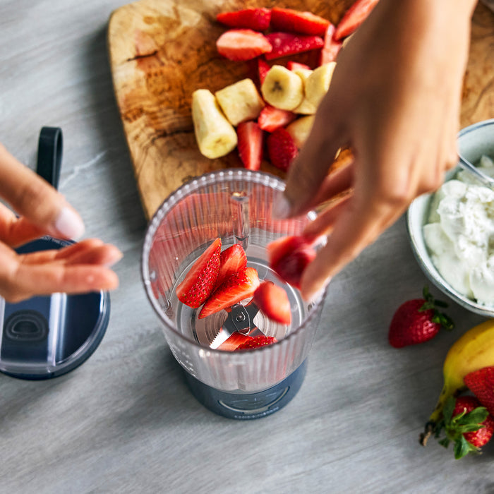 Person preparing ingredients for a smoothie in a blender on a wooden cutting board.