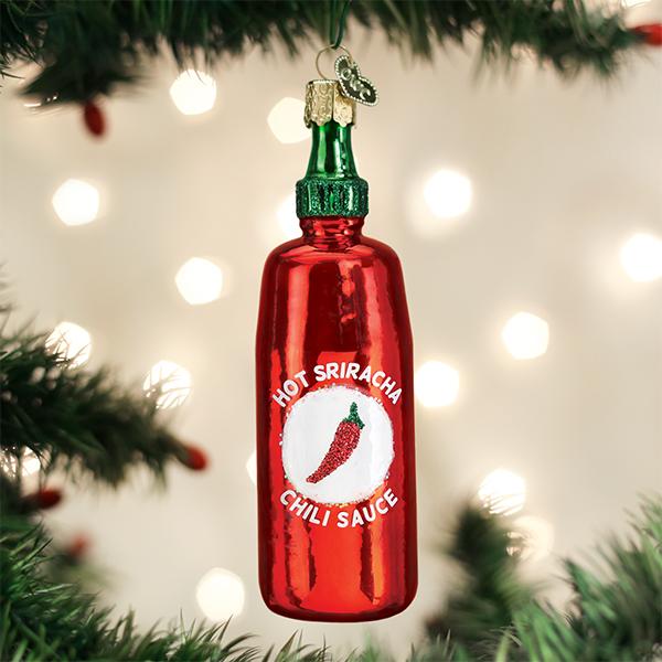 A shiny red Christmas tree ornament shaped like a Sriracha hot sauce bottle hangs from a tree branch, with blurred lights glowing in the background.