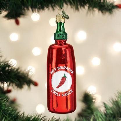 A shiny red Christmas tree ornament shaped like a Sriracha hot sauce bottle hangs from a tree branch, with blurred lights glowing in the background.