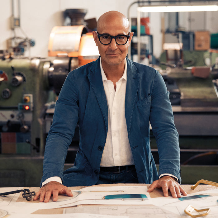 Stanley Tucci in a blue jacket and glasses sitting at a table with blueprints in an industrial setting