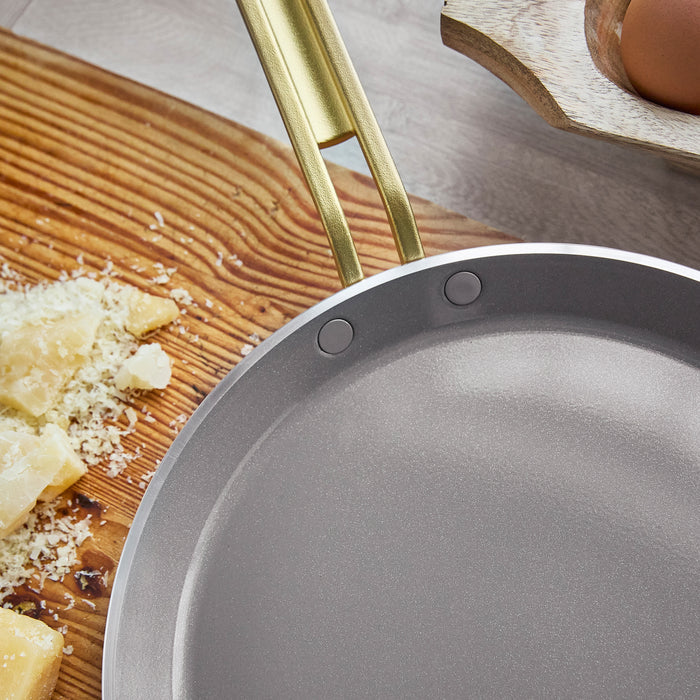 White crepe pan with gold handle on a wooden surface with cheese and eggs.