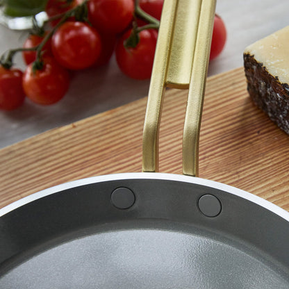 Gold crepe pan handle on a wooden surface with tomatoes and cheese in the background