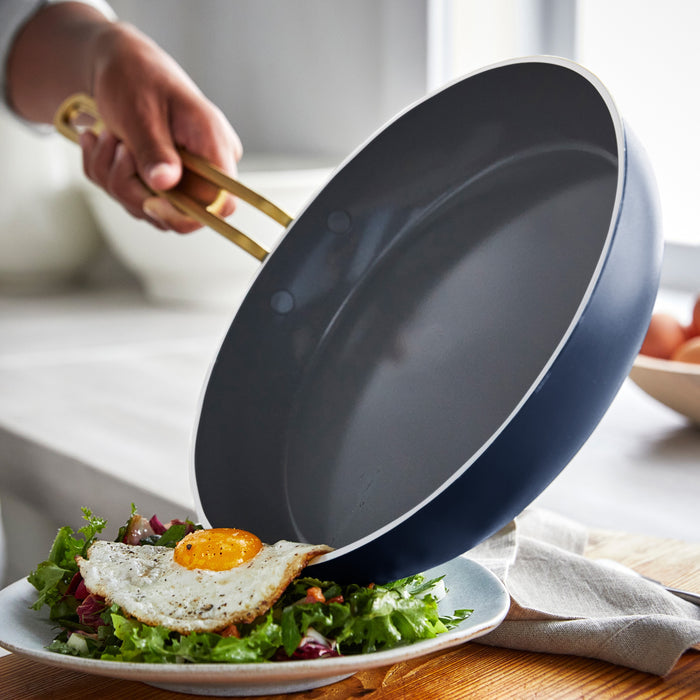 Fried egg being slid from a white Stanley Tucci ceramic nonstick pan onto a salad on a plate.