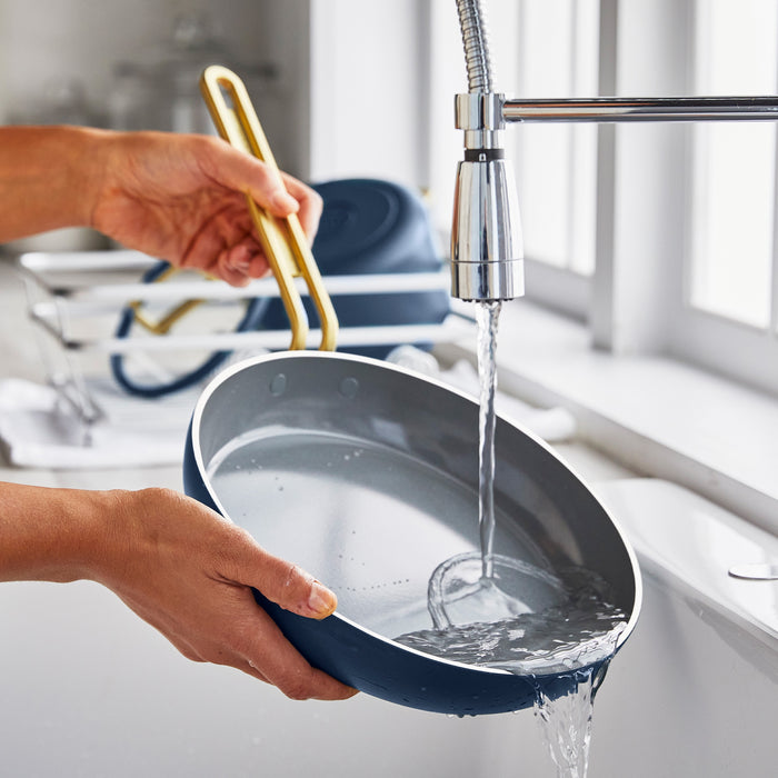 A blue ceramic nonstick mini pan is rinsed under running water in a kitchen sink.