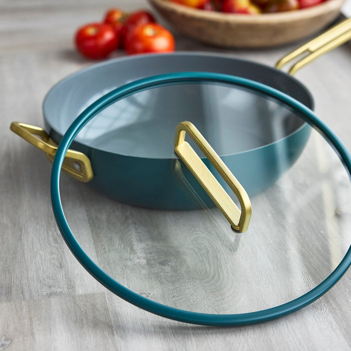 Venetian teal ceramic nonstick pan with glass lid on wood surface; tomatoes in the background.