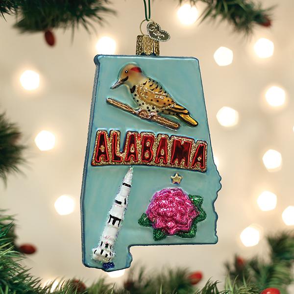 A Christmas ornament shaped like Alabama features a bird, the word â€œALABAMA,â€_x009d_ a rocket, and a pink flower, hanging from a tree branch with festive lights in the background.