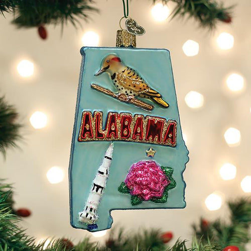 A Christmas ornament shaped like Alabama features a bird, the word â€œALABAMA,â€_x009d_ a rocket, and a pink flower, hanging from a tree branch with festive lights in the background.