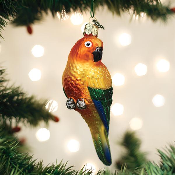 A colorful parrot-shaped ornament hangs from a Christmas tree branch with blurred white holiday lights in the background.