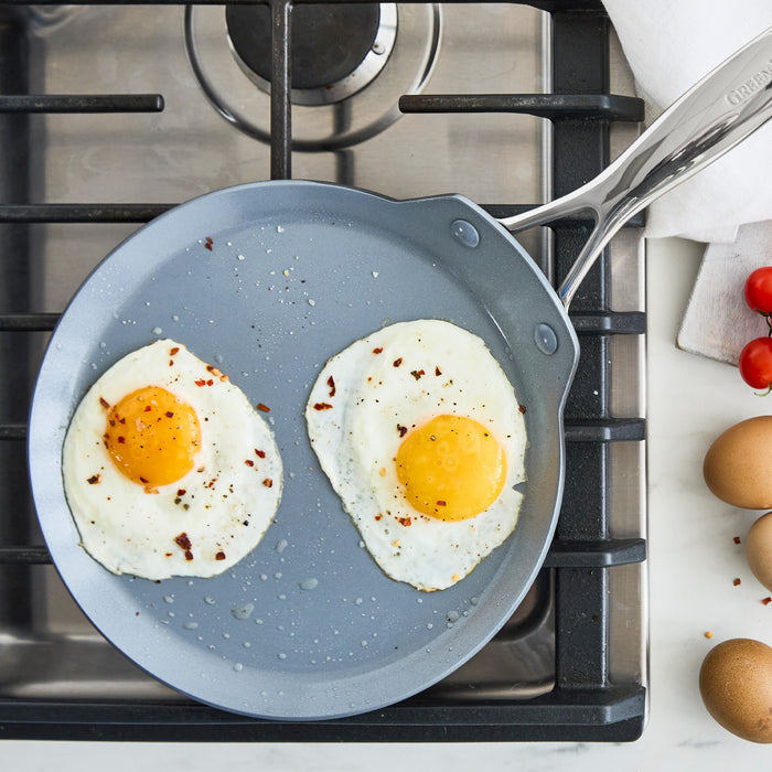 Two sunny-side-up eggs cooking in a gray ceramic nonstick crepe pan, with whole eggs and tomatoes beside the pan.