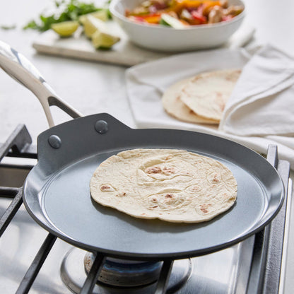 Tortilla on a grey ceramic 9.5 crepe pan on a gas stove, with toppings and tortillas beside the pan.