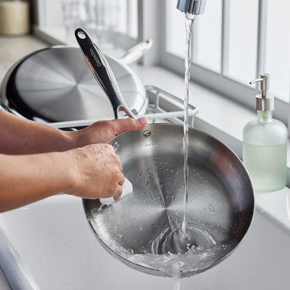 Washing stainless steel frypan set under running water in a kitchen sink, hands visible.