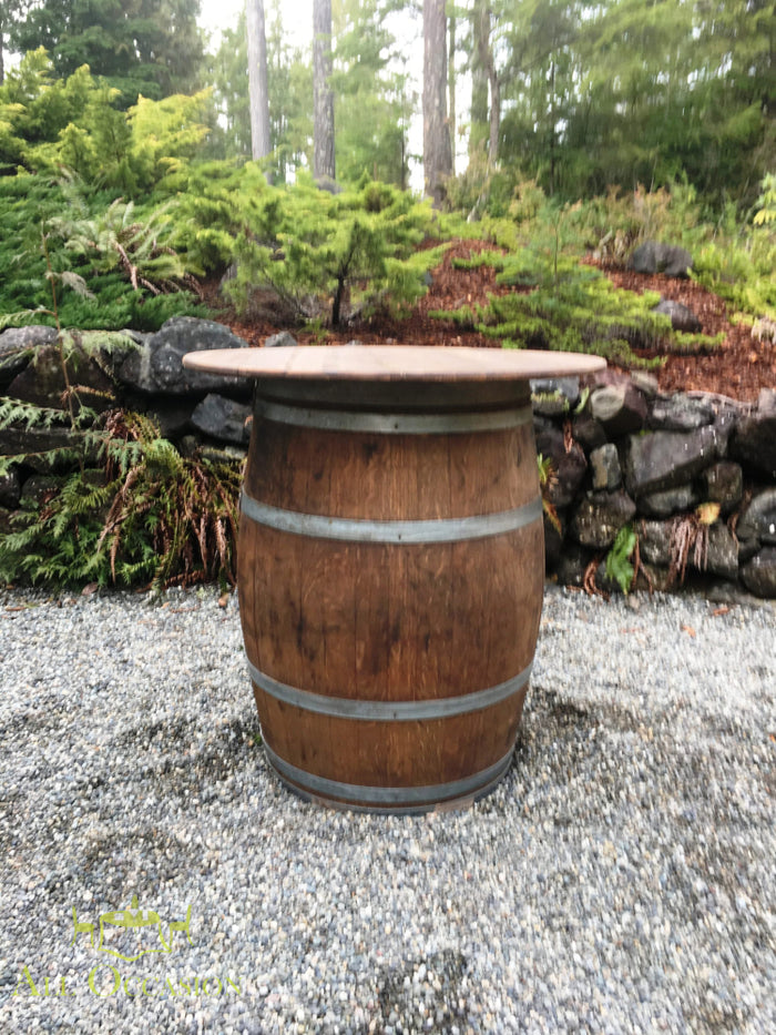 Wine Barrel Cocktail Table Rental, rustic barrel table with wooden top in outdoor setting surrounded by greenery.