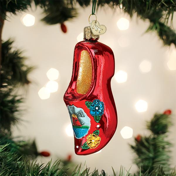 A shiny red ornament shaped like a wooden clog, decorated with colorful flowers and a windmill design, hangs from a Christmas tree branch with blurred lights in the background.